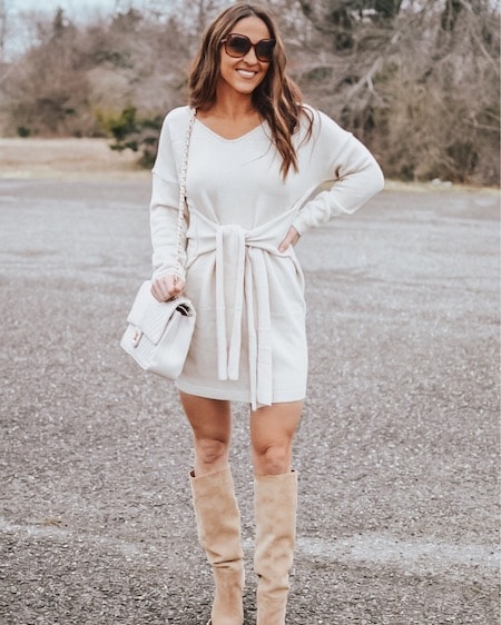A smiling woman in a tie-front sweater dress, tan knee high boots, and brown sunglasses.