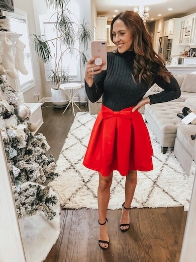 A woman wearing a red mini skirt with a bow, a black sweater, and black ankle strap heels.