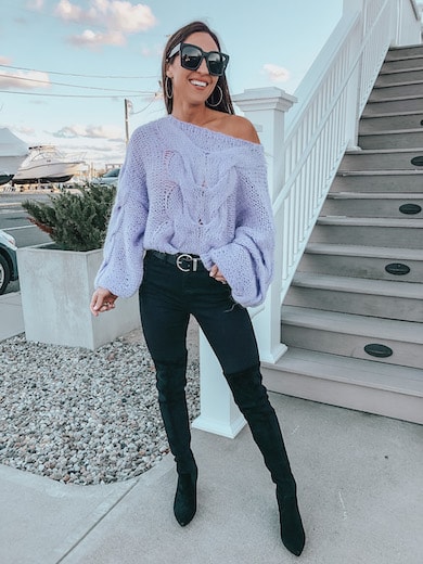A woman wearing a purple sweater and black skinny jeans with black knee high boots.