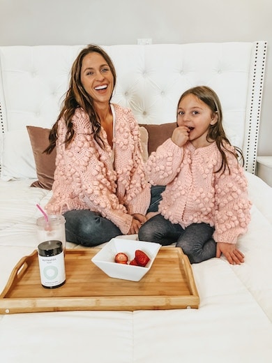 A woman and daughter wearing matching pink cardigans sitting on a bed.