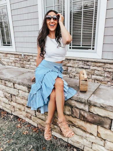 A woman wearing a blue ruffle skirt from Chicwish and a white crop top, sitting on a rock wall.