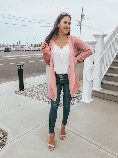 A woman wearing a white tank top, pink cardigan, skinny jeans, and nude ankle strap heels.