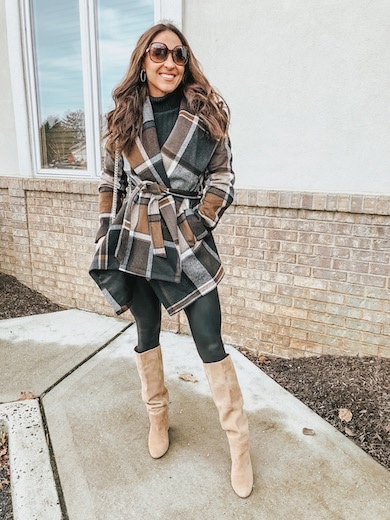 A woman wearing a multicolored plaid wrap coat, black leggings, and tan knee high boots.