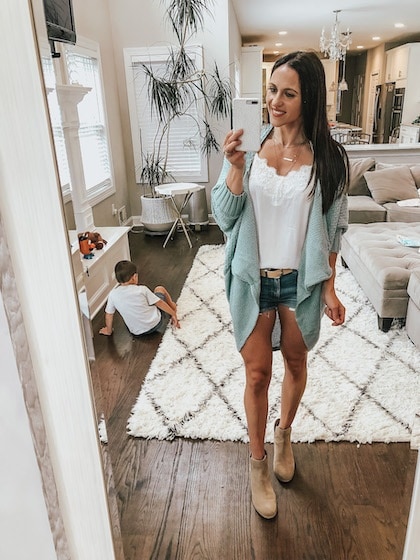 A woman taking a mirror selfie wearing a white tank top, green cardigan, denim shorts, and tan booties.