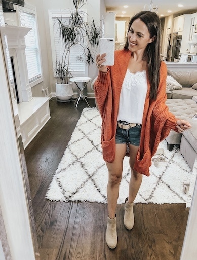 A woman taking a selfie in an orange cardigan and denim shorts.