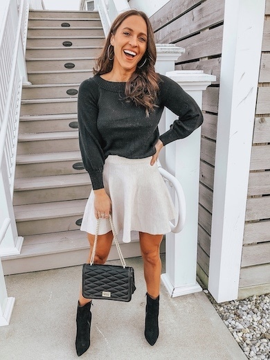 A woman wearing a beige mini skirt, black sweater, and black booties.