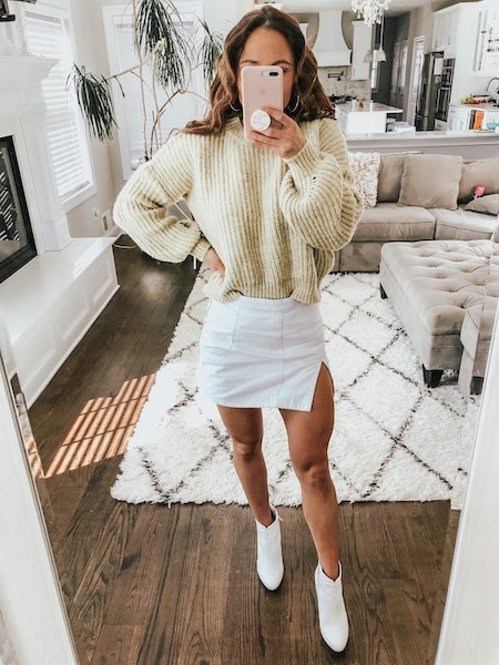 A woman in a yellow sweater and white mini skirt taking a selfie in a mirror.