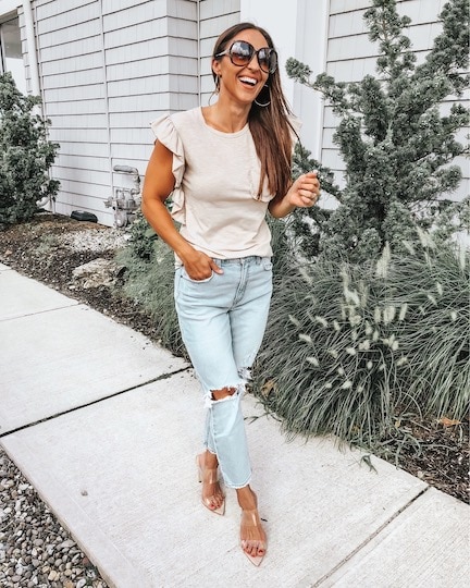 A smiling woman wearing a cream tank top, light wash jeans, and clear heels. 