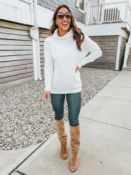 A woman in a white sweater, blue jeans and knee high boots