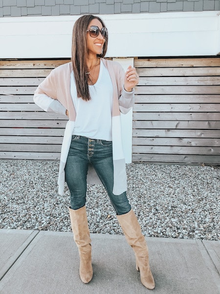 A woman wearing a colorblock cardigan sweater, white tank top, blue jeans, and knee high boots