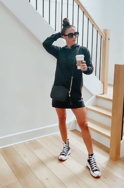 A woman wearing a black sweater dress, black converse sneakers, and black sunglasses.