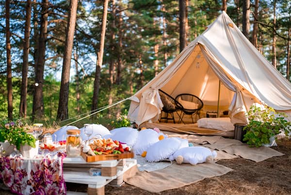 A glamping tent at a campsite with a picnic table, pillows, and rugs outside,