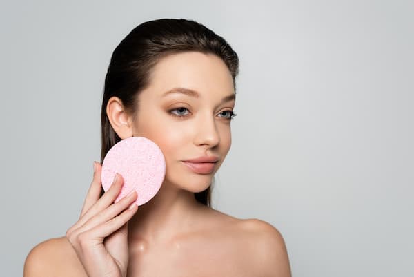 A woman exfoliating her face with a pink sponge.