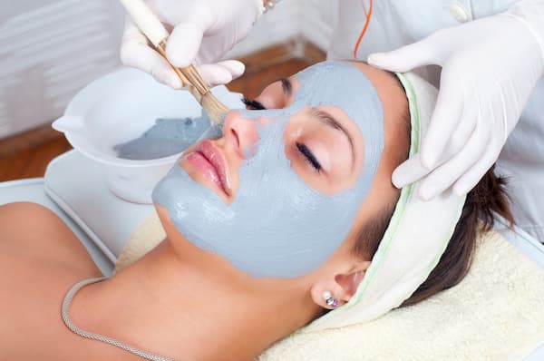 A woman having a blue face mask put on during a facial treatment.