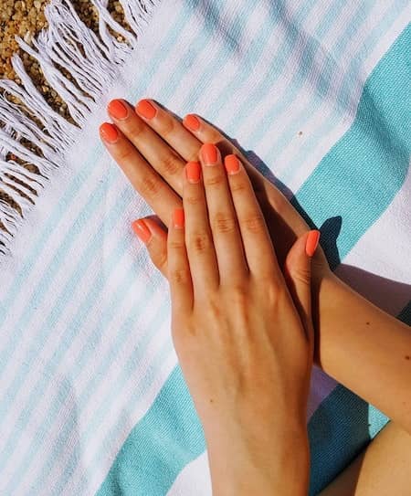 A woman with SNS nails in an orange color holding her hand over a blue and white beach towel.