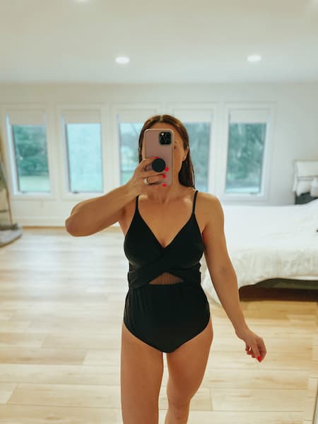 A woman taking a mirror selfie wearing a mesh black one piece bathing suit.