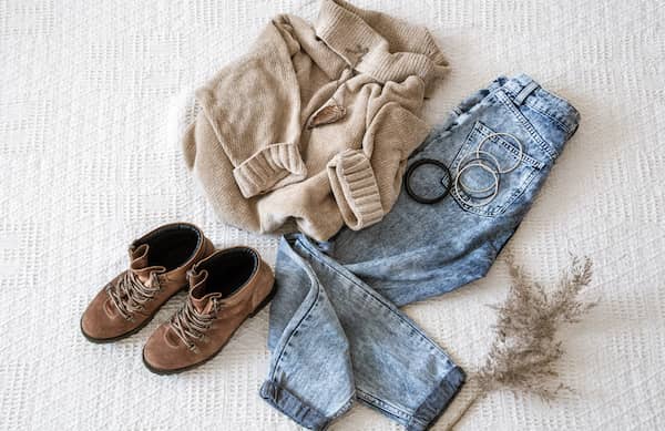 Woman's clothing items on the floor styled together as an outfit including brown boots, a tan sweater, blue jeans, and bracelets.