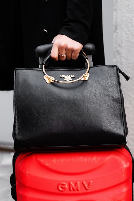 A woman holding a black Prada bag on top of a red suitcase.