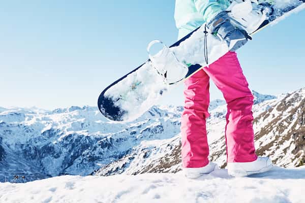 A woman in pink snow pants standing on the top of a snowy mountain holding a snowboard.