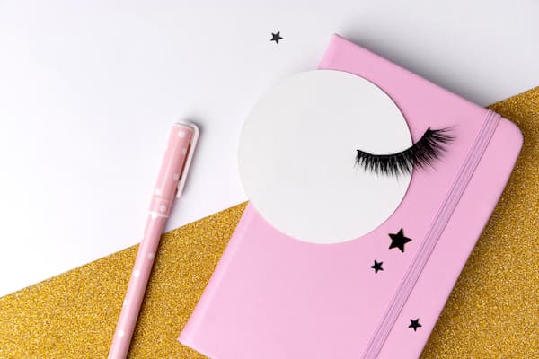 eyelashes on a piece of paper on a pink notebook with a pink pen sitting next to it.
