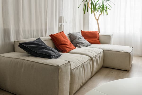 A taupe couch with orange and grey throw pillows on it.