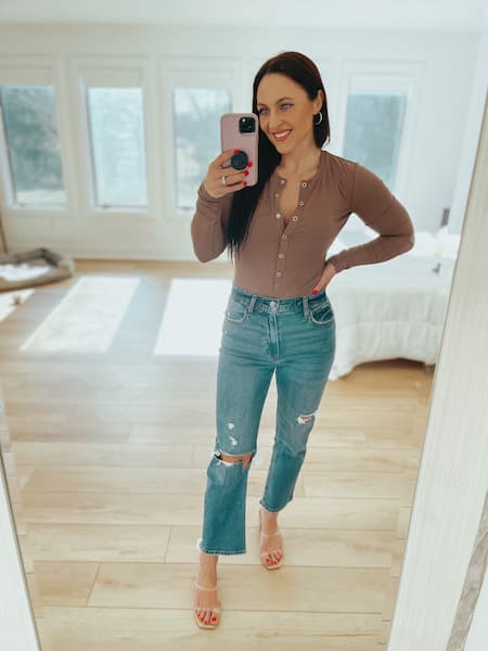 A woman wearing a brown bodysuit, blue jeans, and clear heels taking a mirror selfie.