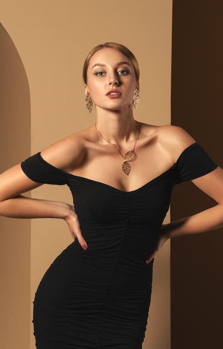 A blonde woman wearing a black off the shoulder dress and a gold necklace.
