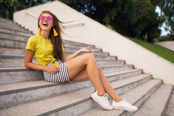 A smiling woman wearing a yellow top, white sneakers, and a striped skirt. 