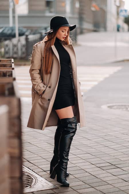 A woman wearing a black dress, a tan coat, a black hat, and black over the knee boots.