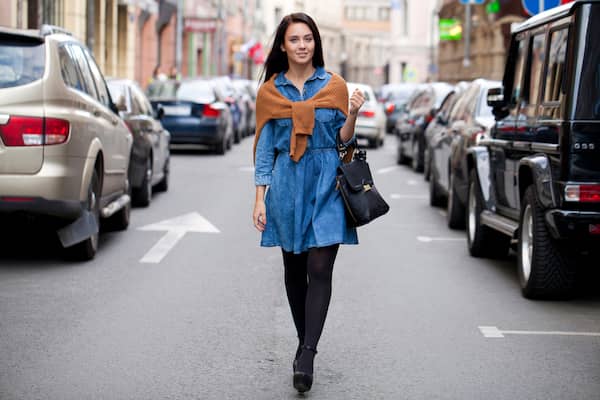 A woman walking in the streets wearing a blue dress, black tights, and a tan sweater over her shoulders. 