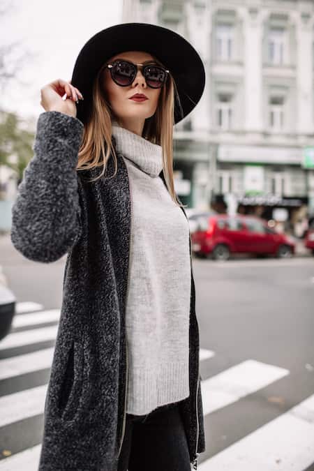 A woman walking in the city wearing a pair of sunglasses, a hat, a sweater, and jacket.