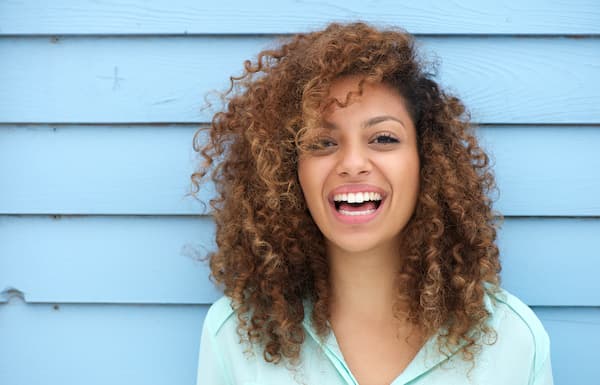 A woman with curly hair smiling against a blue wall.