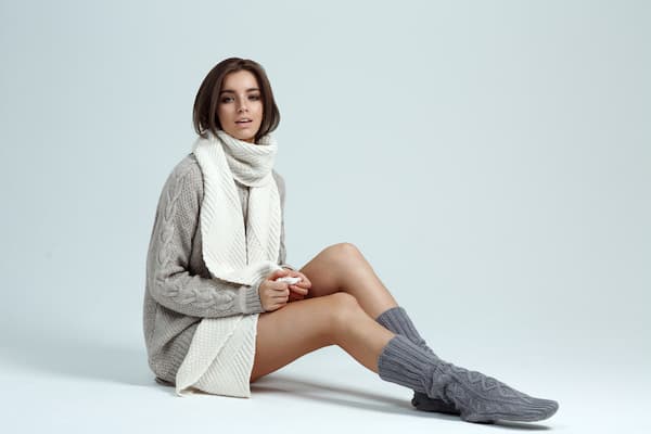 A woman sitting down wearing a sweater dress, scarf, and high socks.