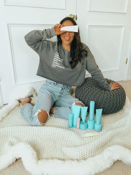 A woman covering her eyes and sitting next to a pouf and some skincare products.
