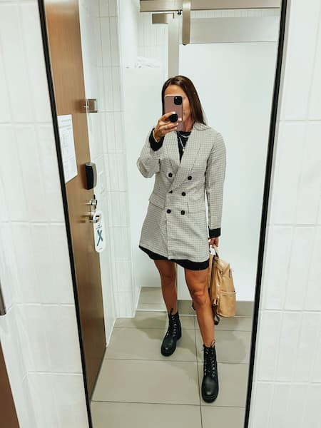 A woman taking a selfie in the mirror wearing black combat boots, a black t-shirt dress, and an oversized blazer.