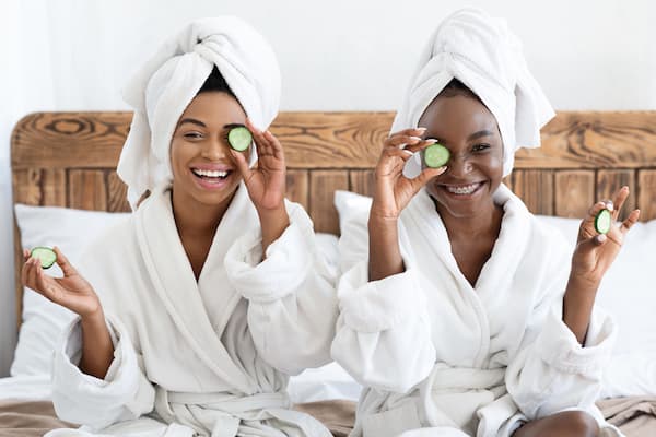 Two girls wearing robes holding cucumbers over their eyes.